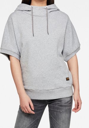 Personne portant un sweat à capuche gris clair à manches courtes et un jean gris foncé déchiré, les mains détendues le long du corps.