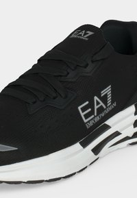 Μαύρο sneaker EA7 Emporio Armani με μαύρα κορδόνια, υφή επάνω μέρος, λευκή ενδιάμεση σόλα και μαύρα γεωμετρικά σχέδια στη σόλα.