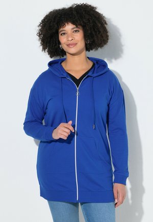 Vrouw met krullend haar, gekleed in een blauwe hoodie met rits over een zwart topje en lichtblauwe jeans, staande tegen een witte muur.