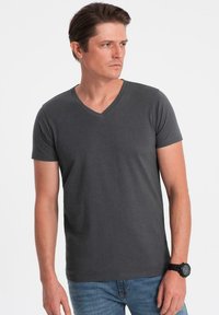 Šedé, krátké tričko s výstřihem do V vyrobené z hladké bavlny. Má fitted design a minimální švy. Nosené s modrými džíny a hodinkami.