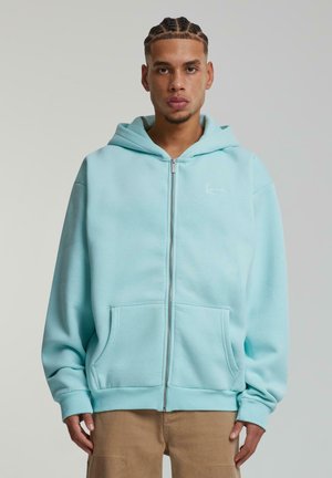 Light blue zip-up hoodie made from soft material, featuring a front pocket, ribbed hem and cuffs, and a small white logo on the chest.