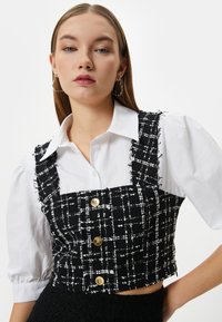 Mustavalkoinen ruudullinen crop-top liivi strukturoidusta kankaasta, jossa on neljä kultanappia, käytetty valkoisen puffy-hihalla varustetun paidan päällä.