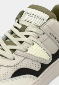 Tenisi albi și verde-oliv, realizati din materiale de plasă și piele. Design perforat, accente contrastante negre și eticheta texturată a brandului pe limbă.