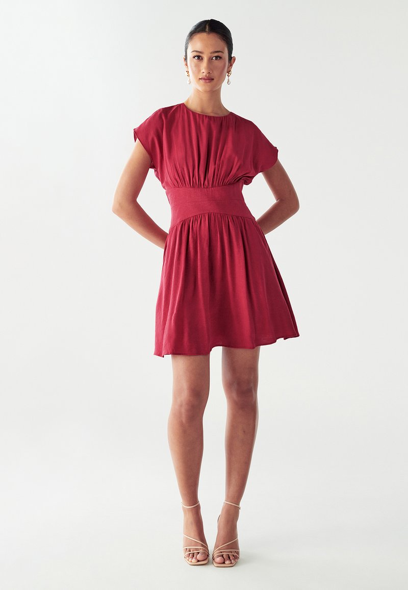 Czerwona sukienka z krótkim rękawem, z marszczonym pasem i rozkloszowanym dołem, wykonana z lekkiego materiału; zestawiona z cielistymi sandałami na obcasie.