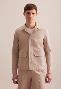 Giacca camicia leggera beige con colletto a punta, chiusura frontale con bottoni, due tasche a toppa e maniche lunghe. Tessuto dalla texture liscia.