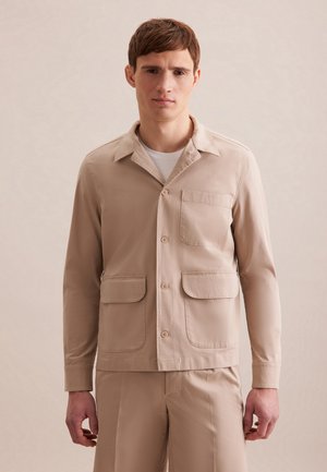 Beige lichtgewicht overhemdjack met een puntige kraag, sluiting met knopen aan de voorkant, twee opgestikte zakken en lange mouwen. Glad stofoppervlak.