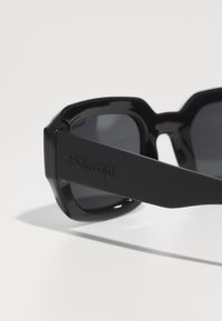 Ochelari de soare negri cu un cadru masiv și unghiular. Brațele sunt texturate și au logo-ul embosat "Polaroid". Lentile întunecate.