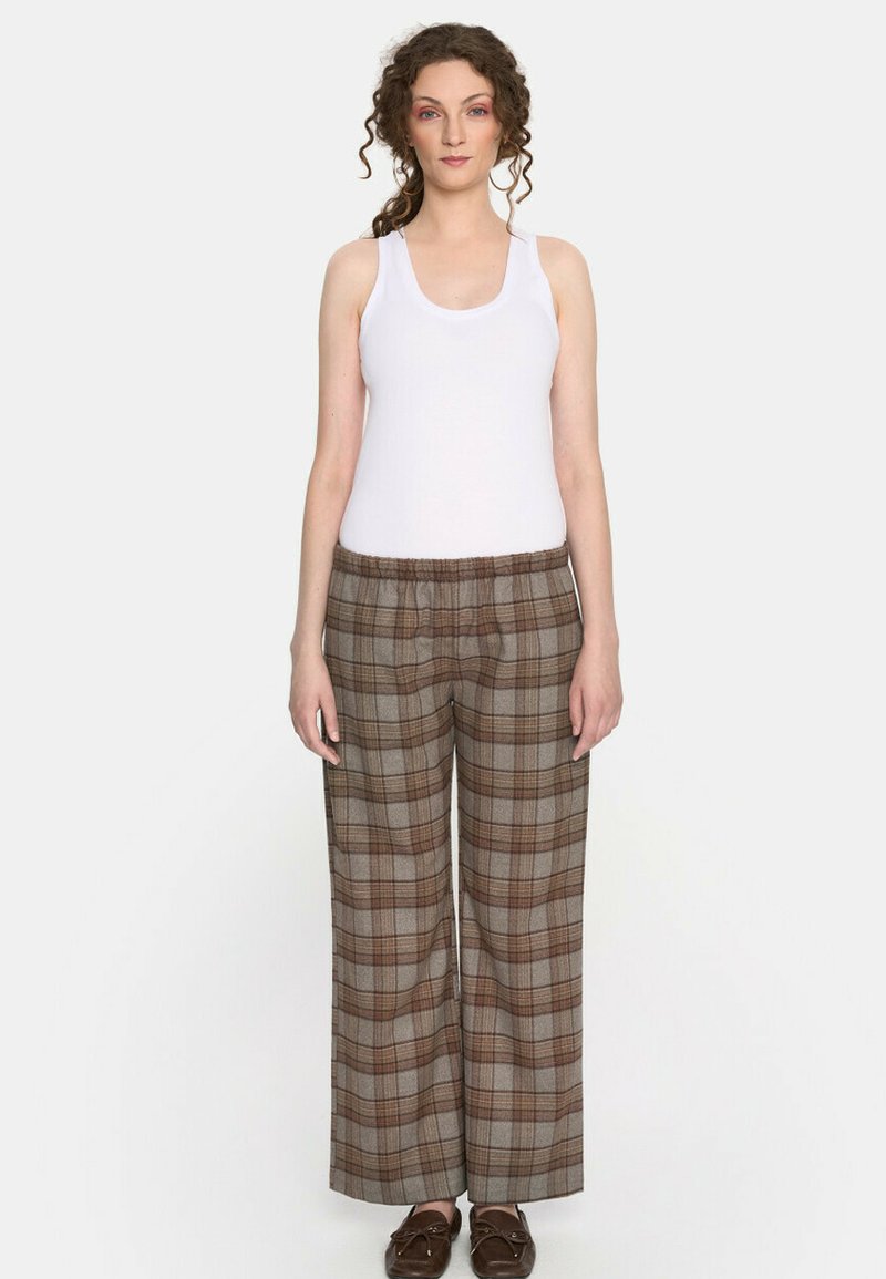 Kobieta z kręconymi włosami, ubrana w białą koszulkę na ramiączkach, brązowe szerokie spodnie w kratę oraz brązowe mokasyny, stojąca na tle jednolitego tła.