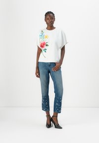 White top with floral design, blue denim jeans with embroidered accents, black high-heeled shoes, displayed against a plain background.
