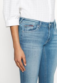 Jeans de denim azul claro con corte recto, bolsillos estándar y una pequeña etiqueta roja y blanca en la parte frontal. Combinado con una camisa blanca de textura.