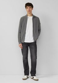 Grijze sweater met rits, witte T-shirt daaronder, donkere spijkerbroek en witte sneakers met bruine accenten. Neutrale achtergrond. Strak ontwerp.