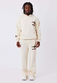 Set di felpa e pantaloni tuta beige con motivi di aerei marroni, realizzato in tessuto morbido. Presenta una tasca frontale e maniche e caviglie elasticizzate.