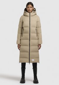 Frau in einem langen beige Puffercoat mit Kapuze, schwarzen Reißverschlussdetails, schwarzen Leggings und schwarzen Schnürstiefeln, die vor einem schlichten Hintergrund steht.