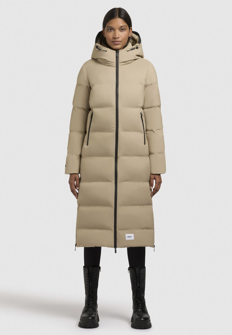 Frau in einem langen beige Puffercoat mit Kapuze, schwarzen Reißverschlussdetails, schwarzen Leggings und schwarzen Schnürstiefeln, die vor einem schlichten Hintergrund steht.
