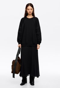 Sudadera negra con acentos de tachuelas en las mangas, combinada con una larga falda negra plisada. El modelo lleva un bolso marrón con una correa. Botas negras.