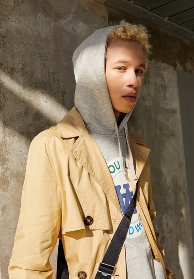 Beige trenchcoat med knapdetaljer over en grå hoodie med grafisk tekst, båret med en sort skulderrem tværs over brystet.