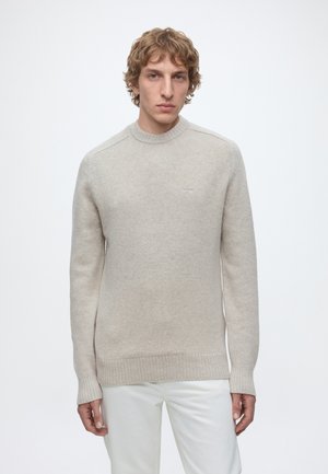 Jeune homme aux cheveux bouclés châtain clair, portant un pull en tricot beige et un pantalon blanc sur un fond blanc uni.