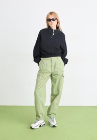 Schwarze, Reißverschluss-Sweatjacke mit hohem Kragen, kombiniert mit hellgrünen Cargo-Hosen mit Seitentaschen. Weiße sportliche Sneaker runden das Outfit ab.