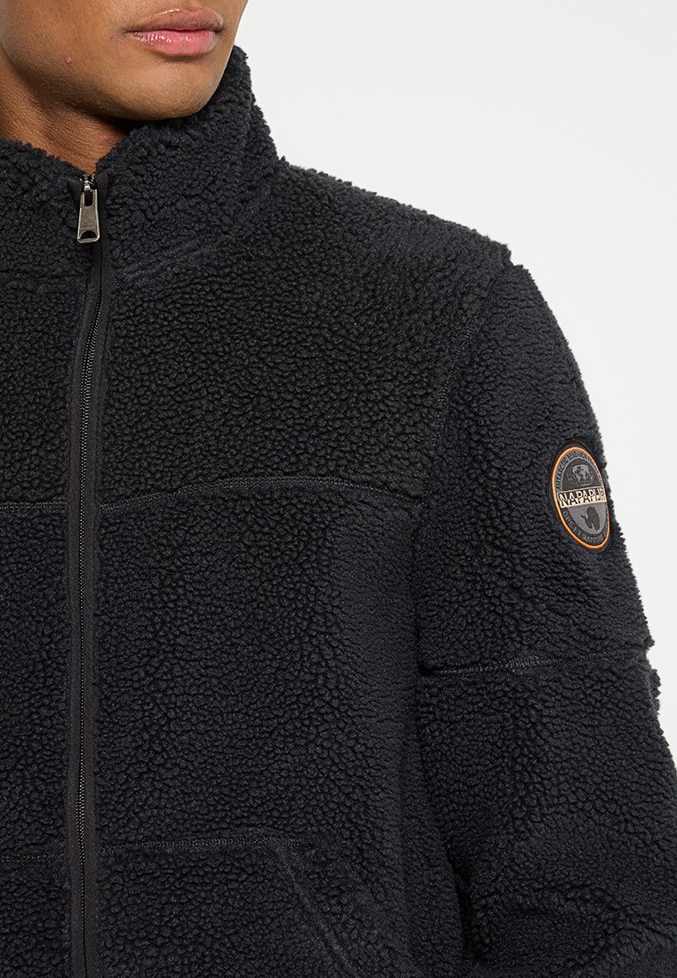 Schwarze Fleecejacke mit weicher, strukturierter Oberfläche, hohem Kragen und Frontreißverschluss. Verfügt über ein rundes Logopatch auf dem linken Ärmel.