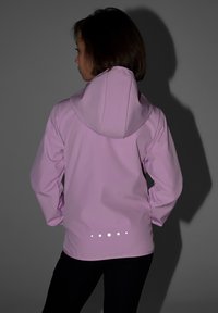 Enfant portant une veste à capuche violet clair avec des points réfléchissants sur le bas du dos, debout les mains sur les hanches devant un fond gris.