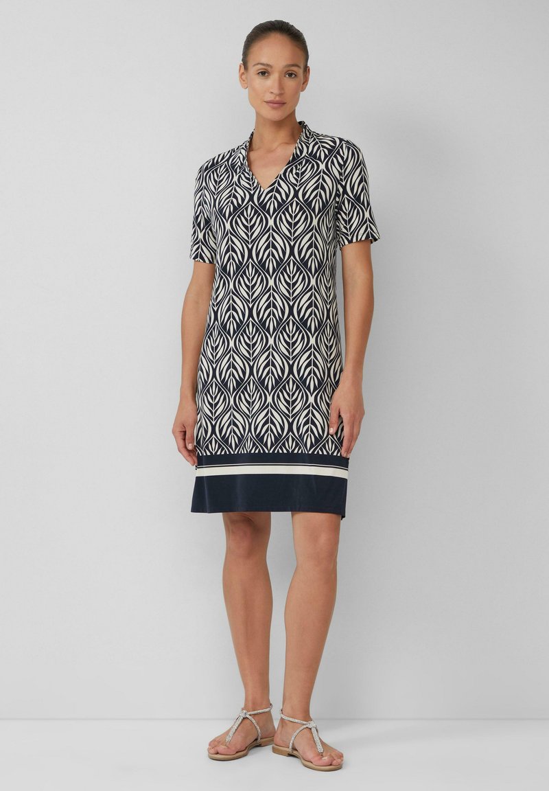 Kurzärmeliges Kleid mit einem navyblauen und cremefarbenen Blättermuster, V-Ausschnitt und einem einfarbigen navyblauen Saum. Das Material wirkt leicht und hat eine glatte Textur.
