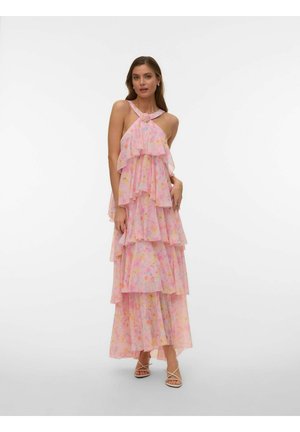 Frau trägt ein ärmelloses, pinkfarbenes, gestuftes Maxikleid mit Blumenmuster, Neckholder und Knotendetail, steht mit einer Hand, die das Kleid berührt.