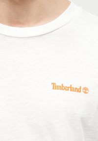 Κλείσιμο πλάνου ενός ατόμου που φοράει λευκό κοντομάνικο μπλουζάκι με στρογγυλή λαιμόκοψη και πορτοκαλί λογότυπο Timberland στο αριστερό μέρος του στήθους.