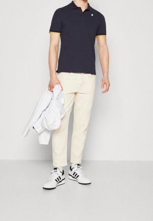 Polo marine avec des accents jaunes, pantalon crème et baskets blanches à rayures noires, tenant une veste blanche. Design minimaliste.
