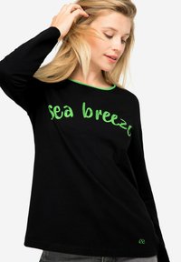 Zwarte langslavende T-shirt van katoen, met groene tekst "zeebries" en een groene kraagaccent. Eenvoudig, casual ontwerp.