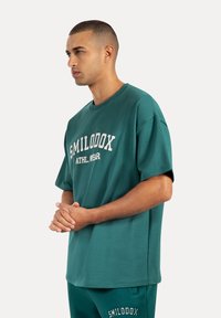 Turkusowa, oversize'owa koszulka z białym napisem "SMILODOX ATHL. WEAR". Wykonana z gładkiego materiału o luźnym kroju.