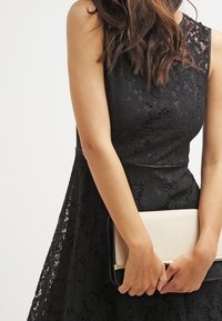 Svart spetsklänning med figursydd bodice och breda band, med en scalloped halsringning. En clutchväska i krämfärg och svart hålls framför.