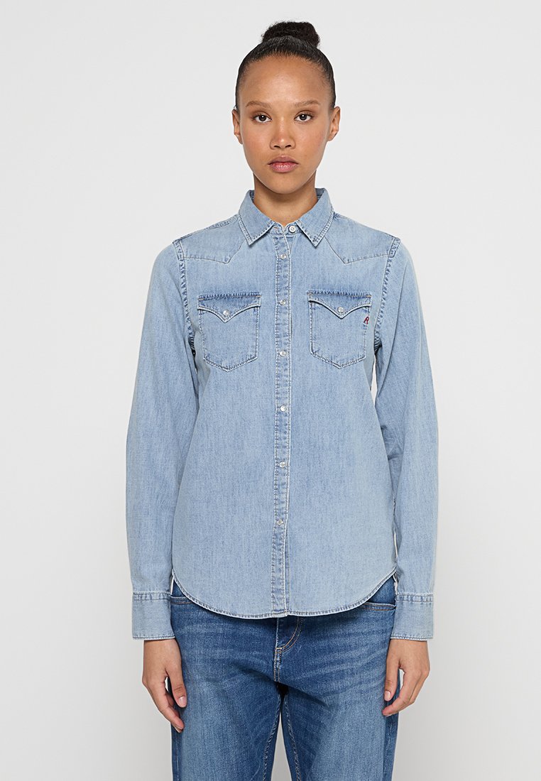 Mujer vestida con una camisa de mezclilla azul claro abotonada con bolsillos frontales y jeans azul medio, de pie contra un fondo blanco liso.