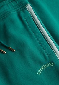 Türkise Jogginghose mit einem Reißverschlussseitenfach, weißem Streifendetail, Kordelzügen mit Metallspitzen und dem "Superdry" Logo, das nahe der Tasche eingestickt ist.