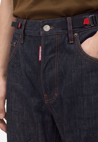Gros plan sur un jean denim bleu foncé avec des surpiqûres orange, une étiquette rouge DSQUARED2, un bouton argenté et une ceinture noire avec des accents rouges.
