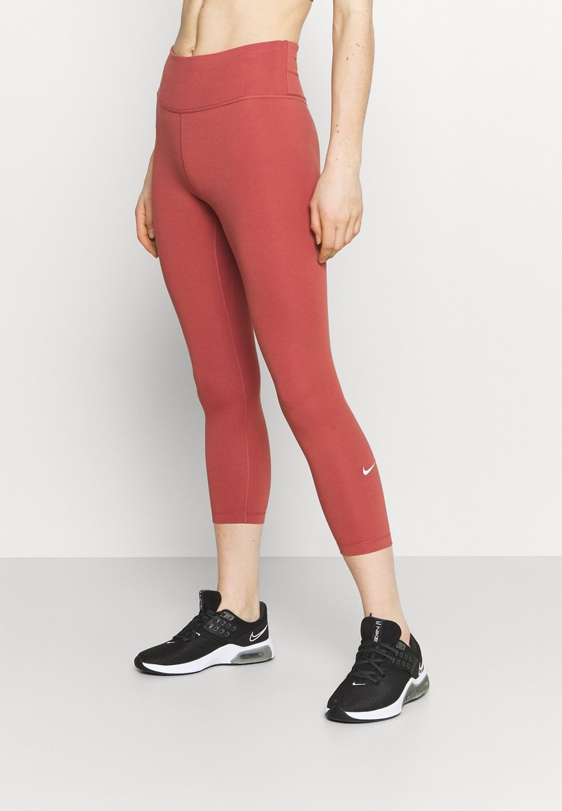 Leggings de sport rouges à taille haute, texture lisse, avec un petit logo Nike blanc près de la cheville. Portés avec des baskets noires.