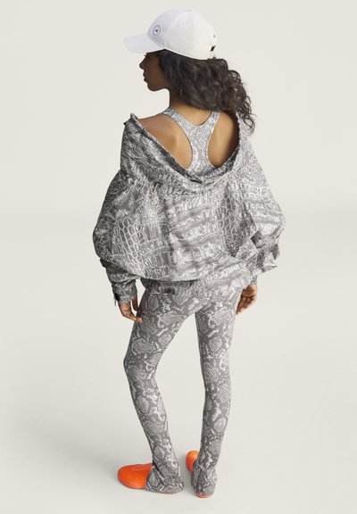 Vrouw die grijze sportkleding met slangenprint draagt, oversized jas die van de schouders is gedrapeerd, witte pet en oranje schoenen, staand tegen een lichte achtergrond.