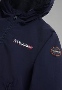 Marineblaue Jacke mit Reißverschluss, Kapuze und Logopatches. Glattes, strukturiertes Material mit orangefarbenen und weißen Akzenten am Branding.