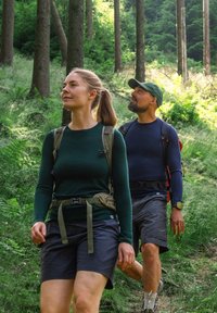 Två personer vandrar på en skogsstig. De har på sig andningsbara friluftskläder: en mörkgrön långärmad tröja, grå shorts och ryggsäckar.