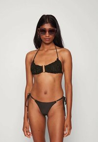 Mulher vestindo um biquíni preto com franzido, com parte de cima tipo halter e parte de baixo com laços laterais, accessoriza com óculos de sol grandes e brincos de argola, standing against uma parede branca.