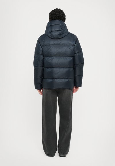 Marineblaue Steppjacke mit Kapuze, ausgestattet mit horizontalen Steppnähten, glatter Textur und elastischen Bündchen. Tragen über grauen Weitbein-Hosen.