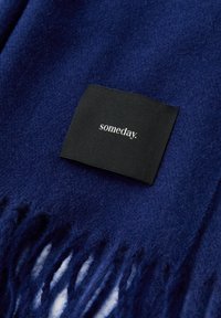 Coperta in pile blu navy con bordi sfrangiati e un'etichetta in tessuto nero con la scritta bianca "someday". Texture morbida e colore uniforme.