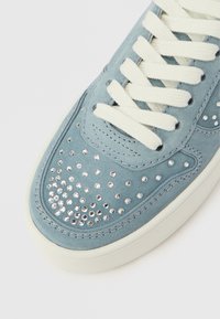 Svetlo modra semišasta superga z belimi vezalkami, okrašena s srebrnimi kristalčki v pikčastem vzorcu na prstnem delu in belo gumijasto podplatom.