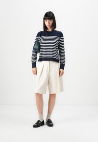 Navy og hvit stripete strikkegenser med skulderknapper, kombinert med kremfargede plissert shorts og svarte loafers. Glatte teksturer og avslappet passform.