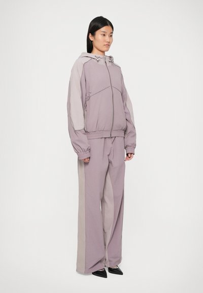 Ανοιχτό μωβ φούτερ με κουκούλα και ελαστικές μανσέτες, συνδυασμένο με χαλαρούς ανοιχτούς μωβ παντελόνες με αντίθετα πλαϊνά πάνελ και τσέπες.