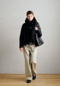 Juodas pūkinis švarkas, per dideli smėlio spalvos kelnių, juoda šalikas su frendžais ir juoda rankinė. Medinė grindų danga ir šviesiai pilka sienos fone.