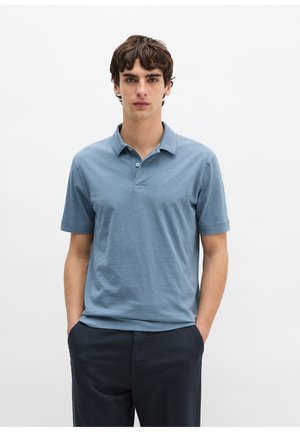 Junger Mann mit kurzem braunem Haar, der ein blaues Kurzarm-Poloshirt und dunkle Hosen trägt, steht mit den Händen in den Taschen.