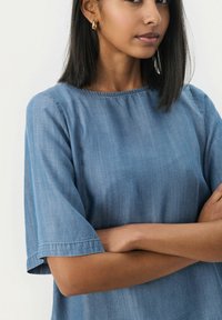 Modra chambray bluza s kratkimi rokavi, okroglim izrezom, subtilno teksturo in sproščenim krojem, poudarjena s šivanim robom.