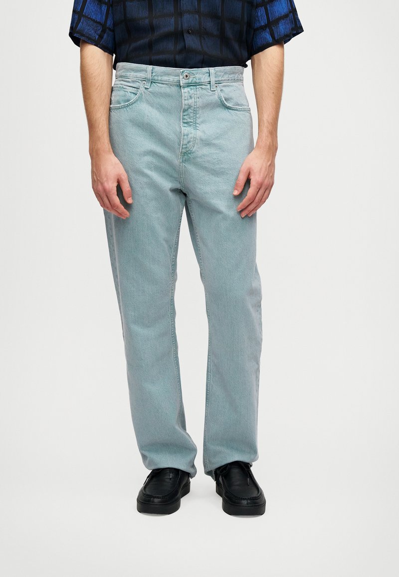 Blugi din denim albastru deschis cu croială dreaptă, având un design clasic cu cinci buzunare și o textură subtilă. Încălțați cu pantofi negri.