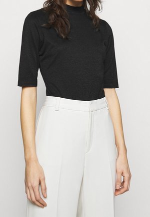 Nainen pukeutuneena mustaan lyhythihaiseen mock neck -paitaan, joka on laitettu valkoisiin korkeavyötäröisiin räätälöityihin housuihin, neutraalia taustaa vasten.