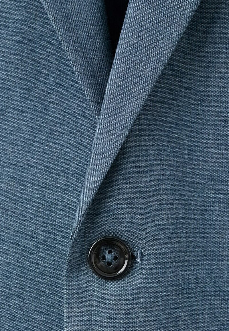 Primer plano de una chaqueta de traje de tela azul con un botón negro de cuatro orificios abrochado cerca del borde de la solapa.